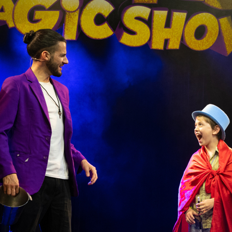 Adult performer pulling a face at a child volunteer dressed as a magician, onstage during the show.