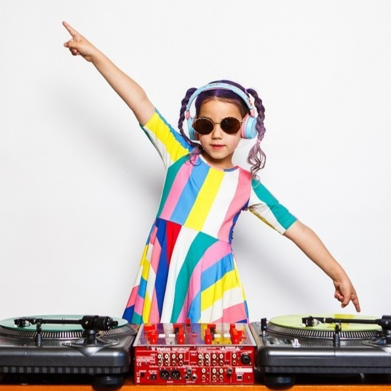 A kid in a colourful dress with purple pigtails is spinning some vinyl records on some decks