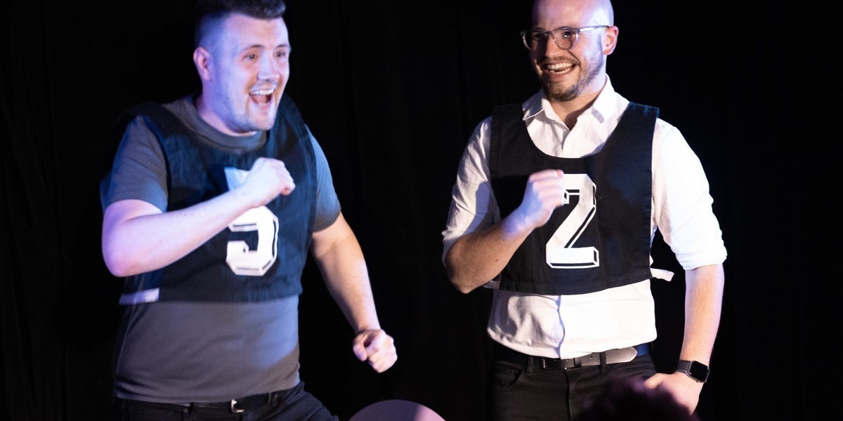Two improvisers performing on stage wearing netball bibs. Each bib has a number on it. The improvisers are smiling and swinging their arms like they are marching or skipping.