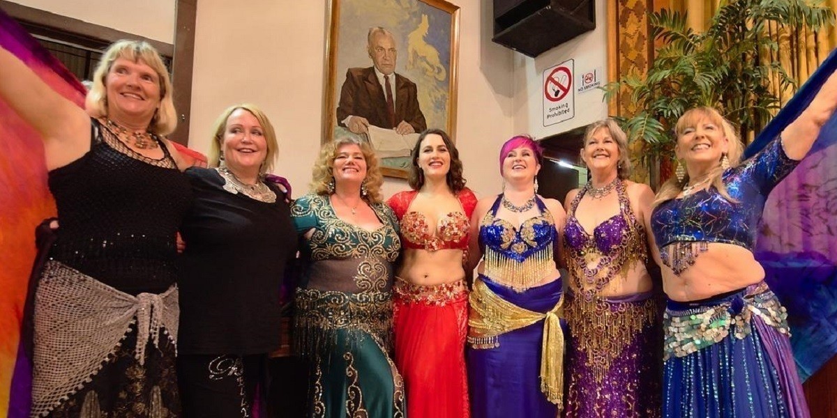 A photograph of seven women in a row smiling for a photo. They are all wearing belly dancing costumes of different colours that are adorned with silver and gold embellishments.