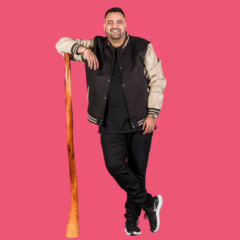 A smiling Aboriginal man wearing black jeans and a brown jacket stands with ankles crossed. He is leaning on a didgeridoo with one elbow.
