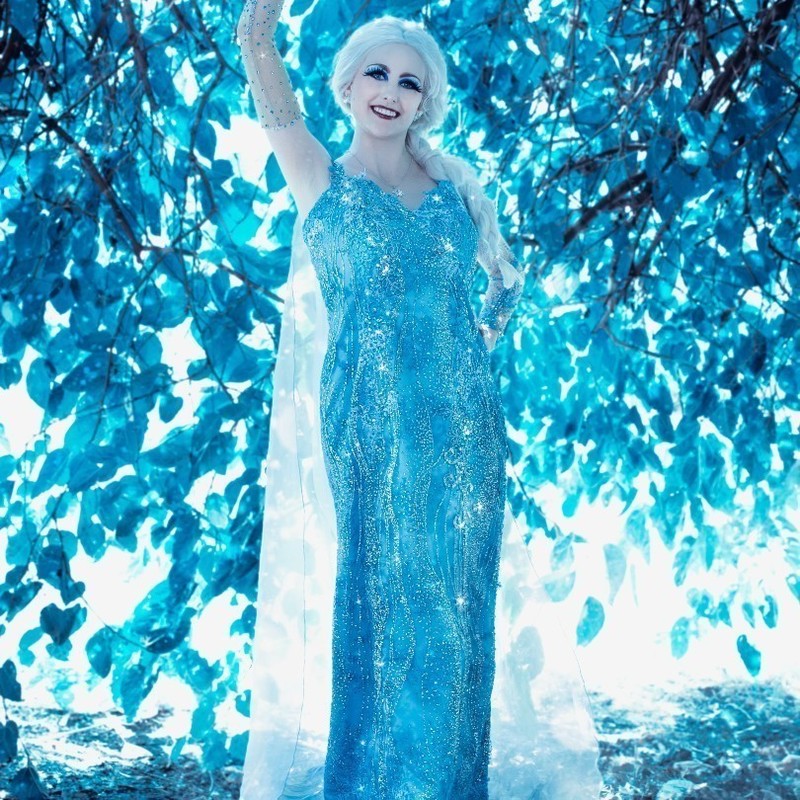 Performer dressed as Disney character Elsa from the movie Frozen. Performer stands with left arm raised in a pose, wearing a long, blue, embellished gown.