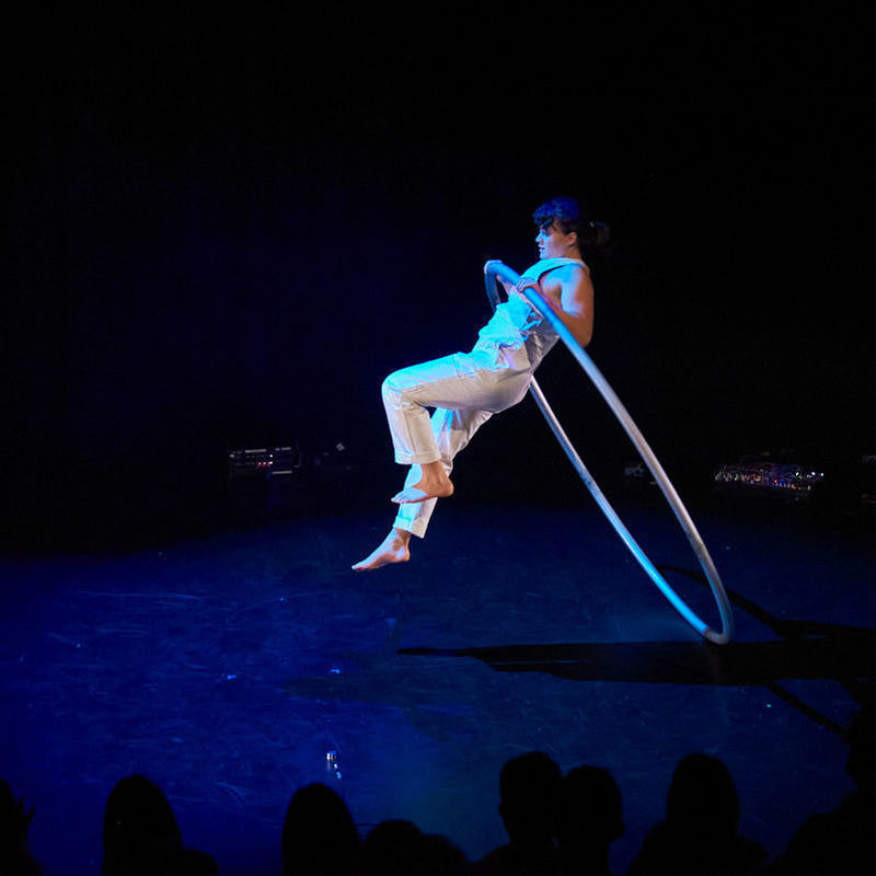 An acrobat wearing white overalls leaps through a cyr wheel, a large metal apparatus, while another acrobat behind them adjusts musical equipment.