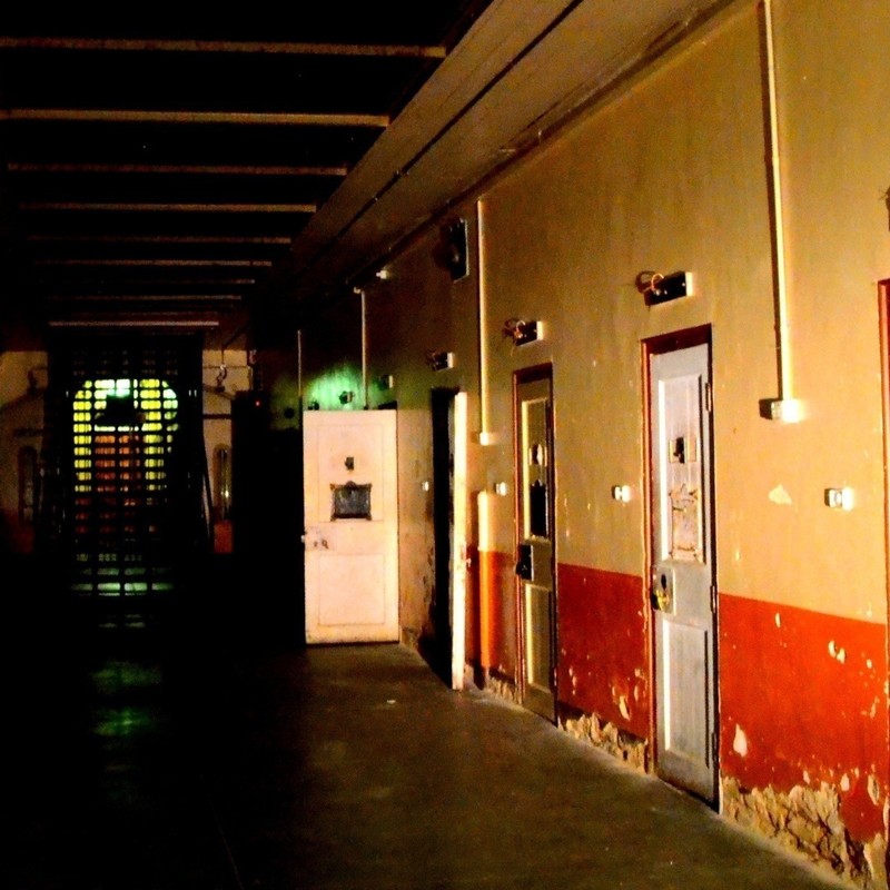 Adelaide Gaol Ghost Tour + Investigation SOUTH AUSTRALIA - A corridor in a cell block with a grey concrete floor. There are three doors along the right hand side of the corridor, the last cell door is open, the wall is painted orange about a third of the way up, the rest is a dirty cream colour. There are chips and holes in the walls and an eerie green light towards the end of the corridor.