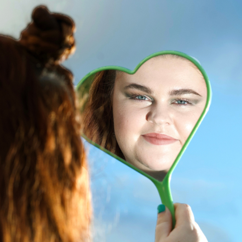 Image of Courtney [lead character and actor] holding a green heart shaped hand-mirror with the reflection of her face central to the camera, against a blue sky.