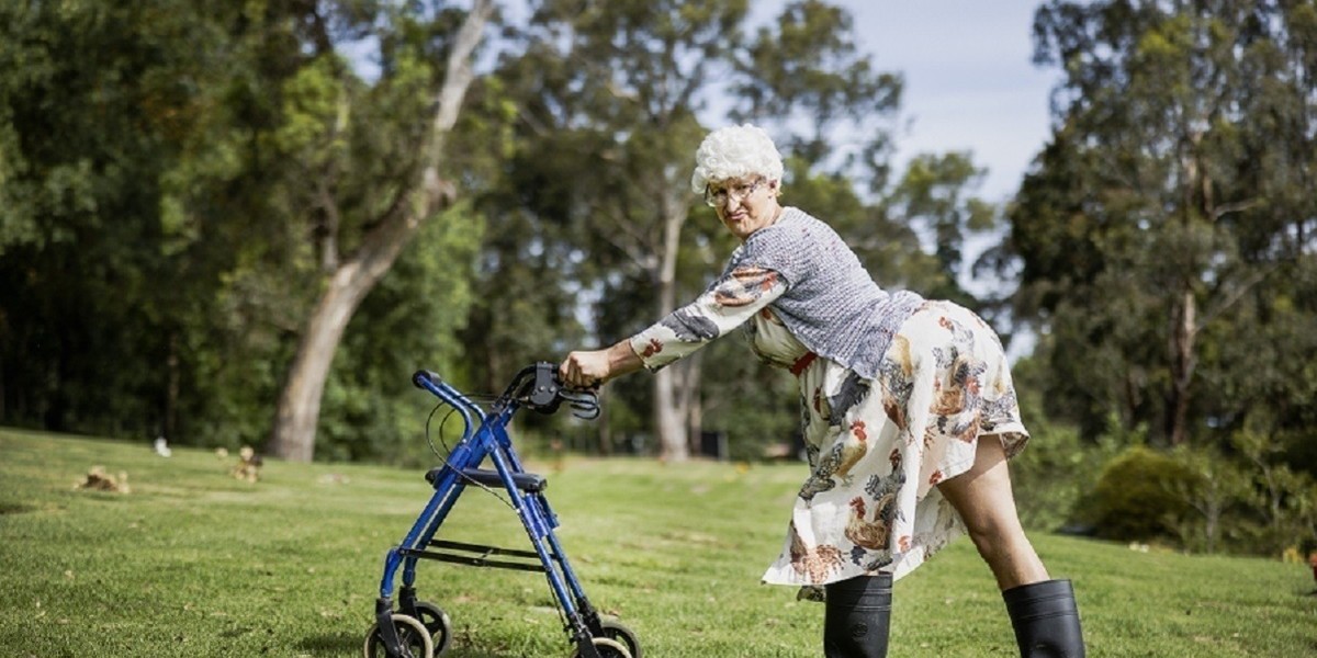 A performer dressed as a granny walking across grass with walker frame in black gum boots