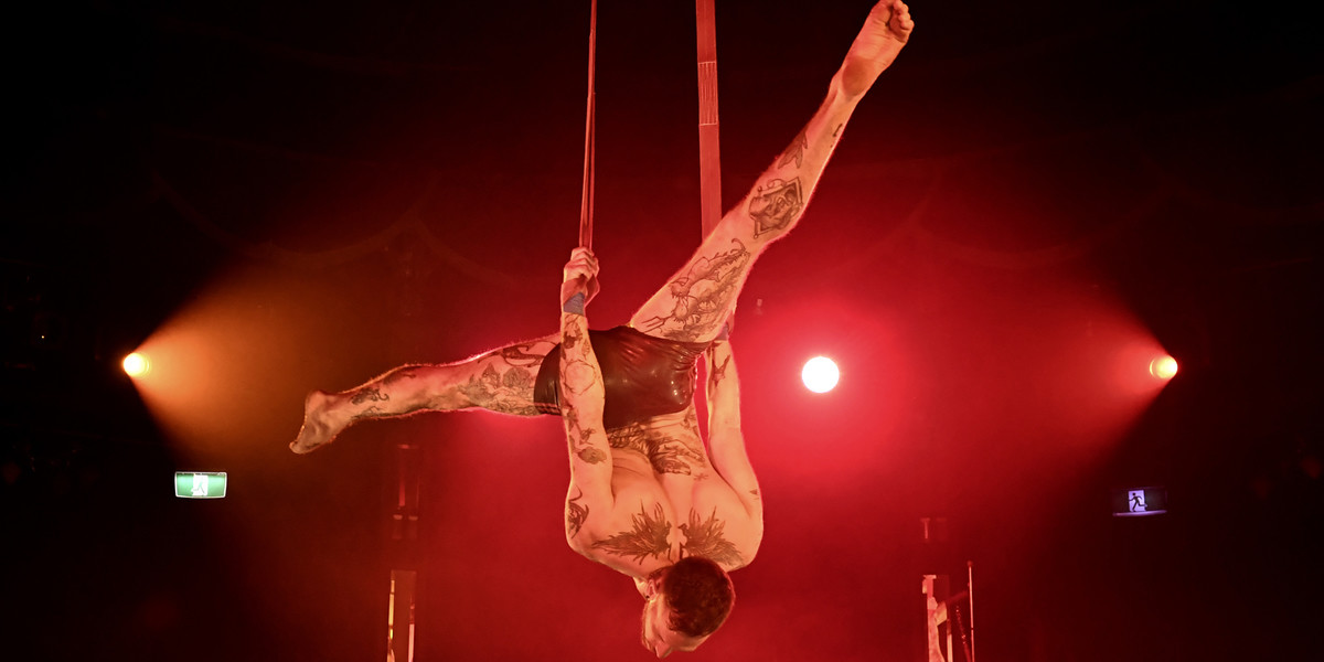 Muscular, tattooed acrobat upside down in the air holding straps and doing the splits