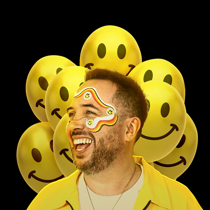 Rob is wearing a bright yellow collared shirt over a white t-shirt. His head is turned to the left, he's smiling broadly with rainbow facepaint design curving across his forehead and down his cheek. Behind him are several graphic representations of yellow smiley face balloons.