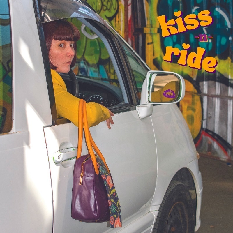 A woman sits inside a white van with a purple handbag hanging out the window. The words 'kiss n ride' appear in the top right corner in yellow text.