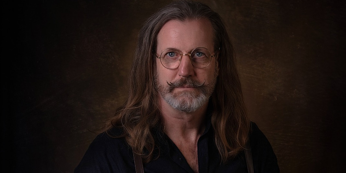 Mister Meredith, a middle-aged caucasian man with long brown hair and a grey beard, is wearing round gold-rimmed glasses and a black shirt with leather braces. You see his head and shoulders facing the camera in front of a sepia background.