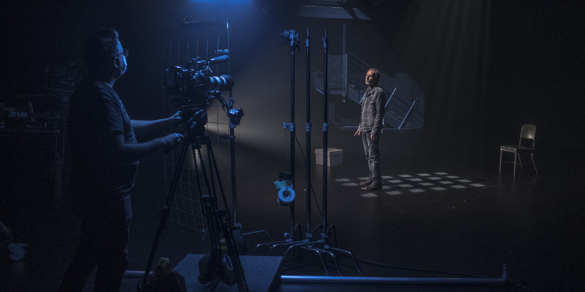 A shot from behind the scenes as a man stands on a dark stage while a camera films him.