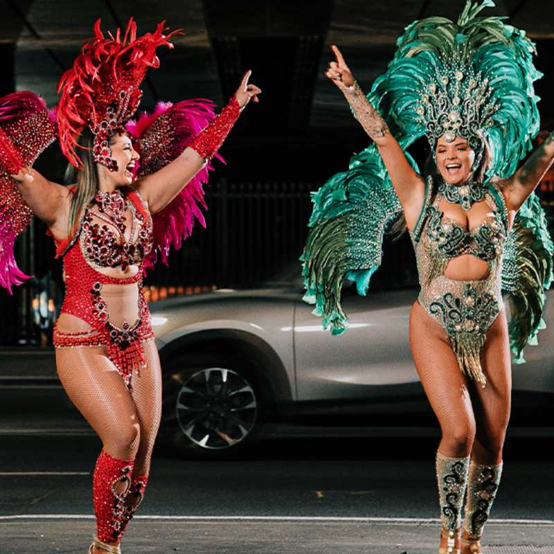 Two women laughing and celebrating with each other on the street, wearing Brazilian carnaval outfits with feathers and sequins.