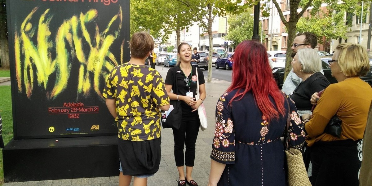A female tour guide speaks to participants about an Adelaide Fringe poster along North Terrace, Adelaide.