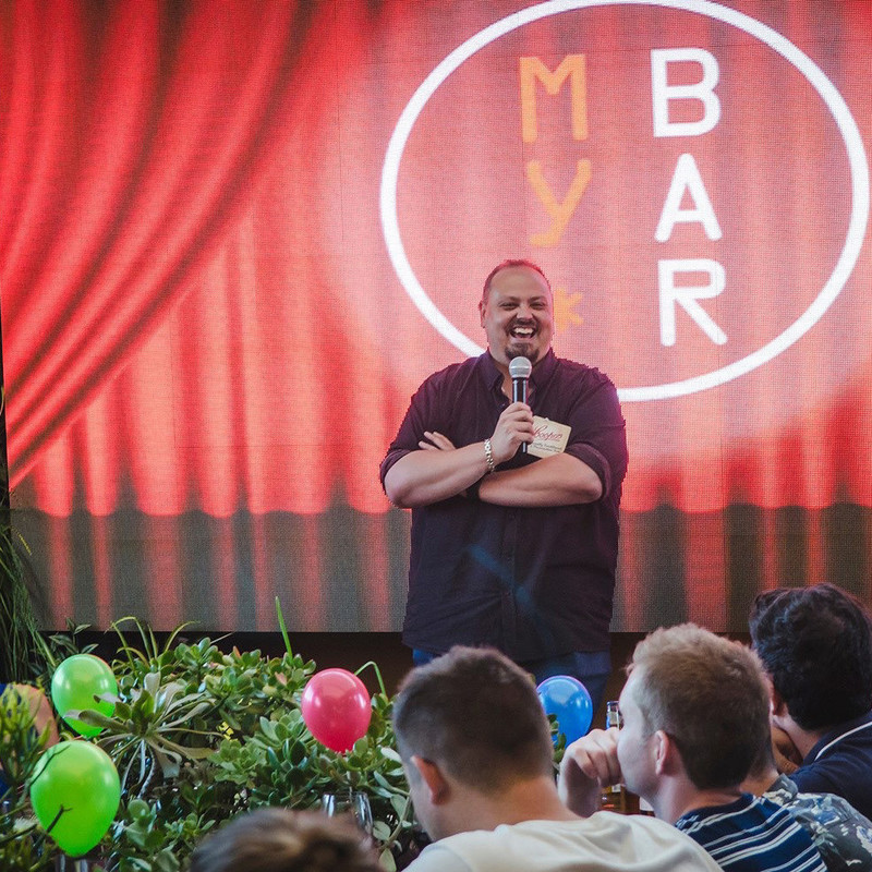 A man stands on a stage with his arms crossed holding a microphone and smiling broadly. The background is a projected red curtain with the text "my bar". The foreground has the heads of an audience and some plants and balloons.