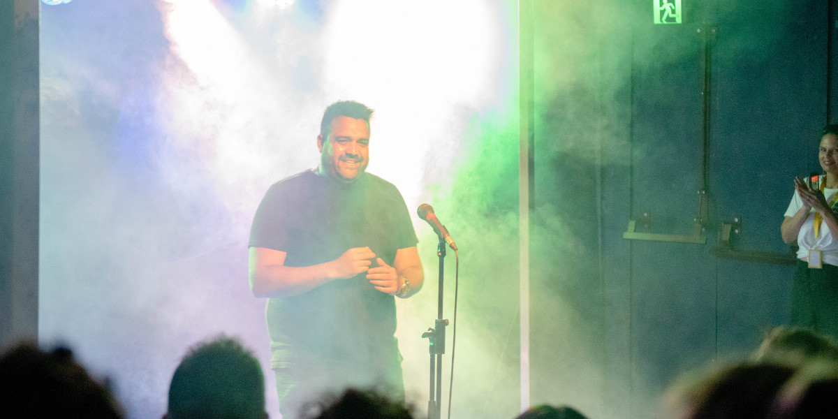 Aboriginal comic Dane Simpson giggles onstage. Green lights and stage haze surround him.