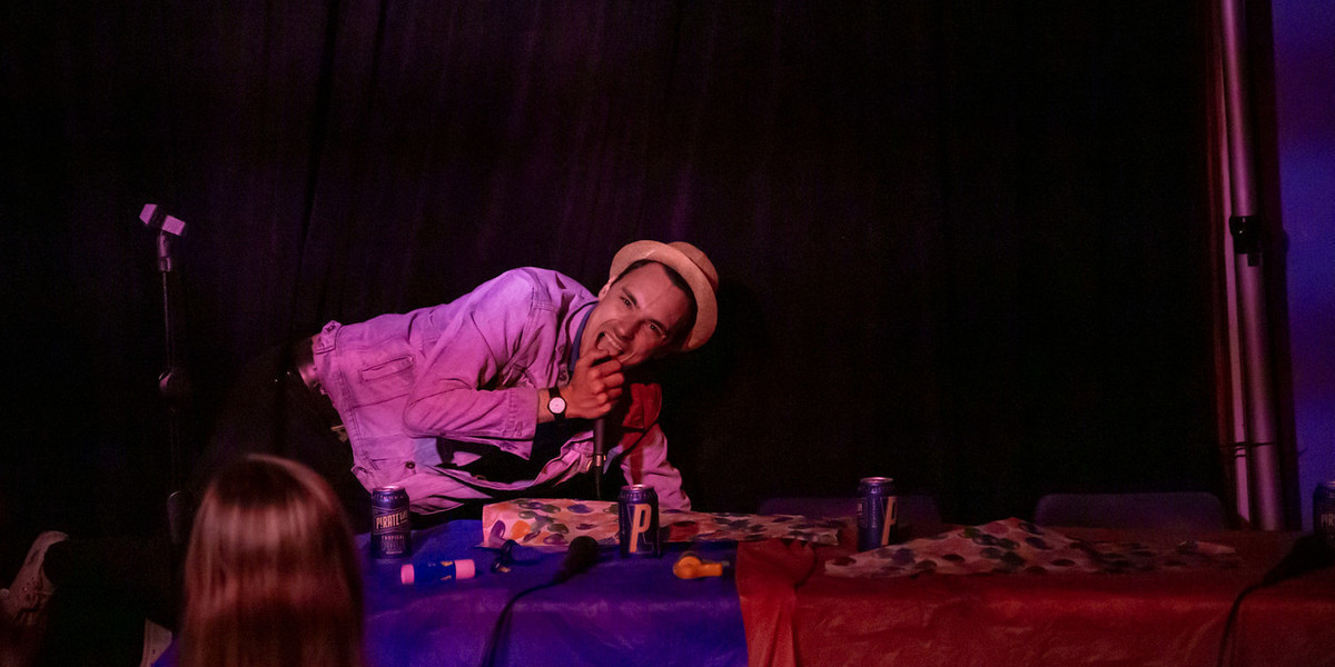 A photo of a man leaning on a table whilst on stage. He is wearing a straw bucket hat and a light coloured long sleeved shirt. He is holding a microphone to his face and has a overjoyed look on his face.