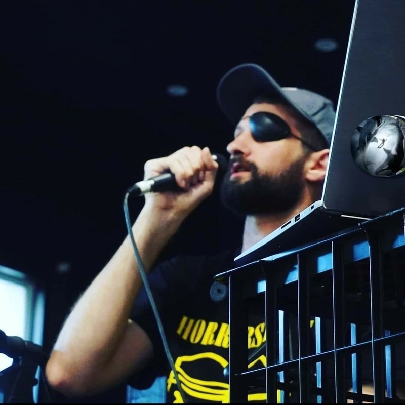 A man stands behind a laptop on a black milk crate. he holds a microphone to his mouth, he is wearing a black t-shirt with yellow writing, a black cap and a black eye-patch.