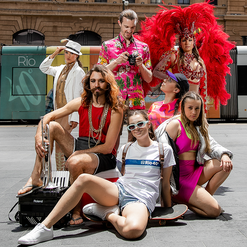 Group is assembled on the pathway
A man stands in Hawaiian shirt holding a camera 
A woman stands holding a hat in a gold dance suit and white suit jacket 
A woman in a bright red carnival feathered suit 
3 people kneel on the ground in dance clothes 
A person with long locks and a fantastic mustache sits on esky holding a trumpet