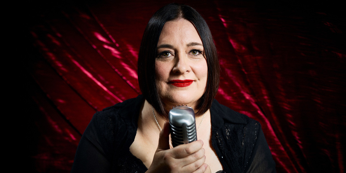 Headshot of Alexandra Frost, Jazz, cabaret singer dressed in black with red velvet background holding vintage microphone
