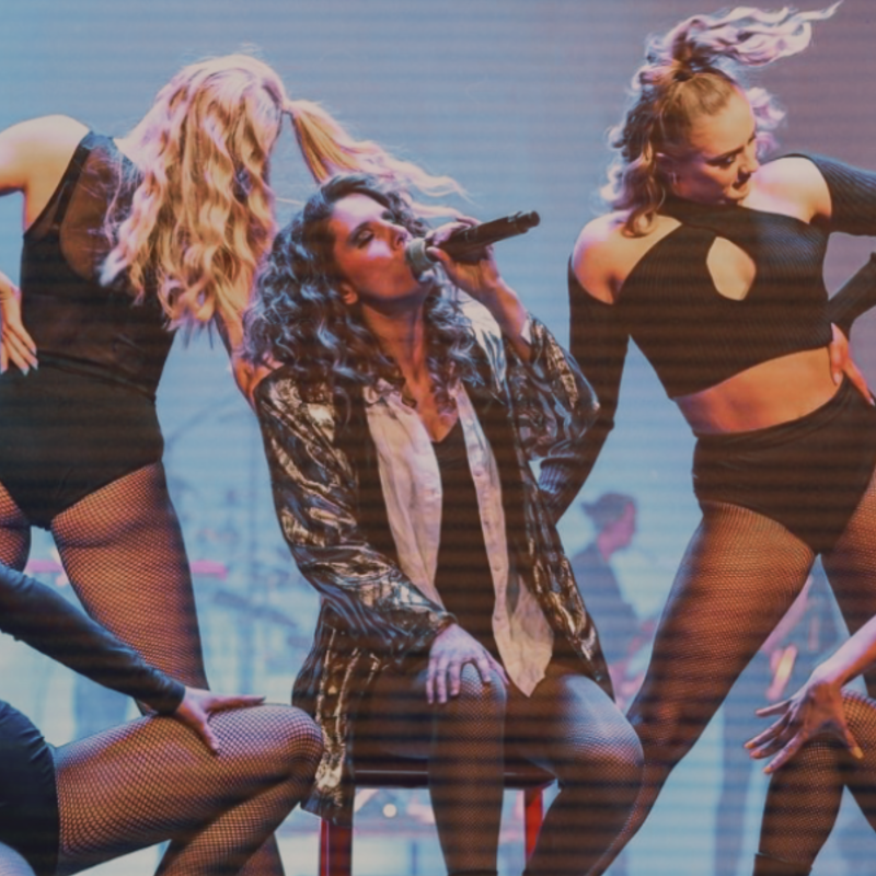 Image of a female performer wearing a gold oversized 80's blazer, sings on a red chair. She is surrounded by 4 dancers wearing black 80's inspired costumes. In the background, you see a string quartet, a saxophonist and a drummer.