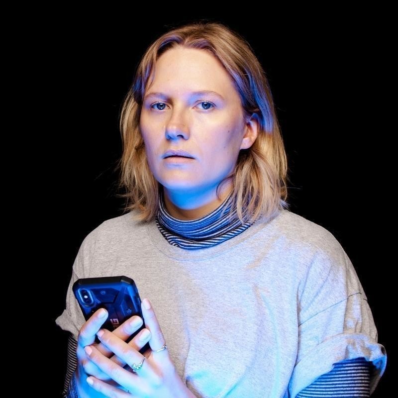 JORDAN BARR - ZOMBIE - Headshot of Jordan Barr looking at the camera, holding an iPhone. Jordan is lit by the phone screen. Black background. She is wearing a grey tee-shirt over a black and white striped turtle neck.