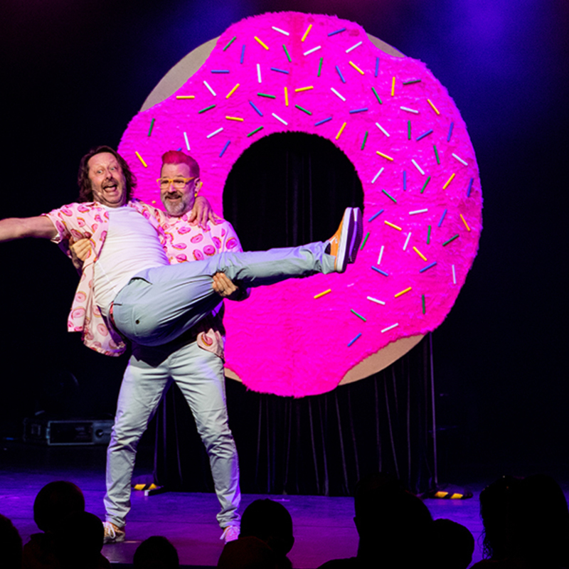 One man in a pink shirt and blue trousers holds another man, also in a pink shirt and blue trousers, caught in his arms — in front of a large pink doughnut.
