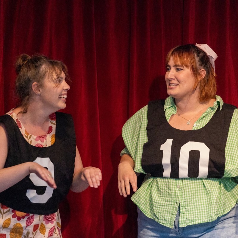 Two female performers wearing numbered sports bibs smile at each other with their hands miming fish flippers.