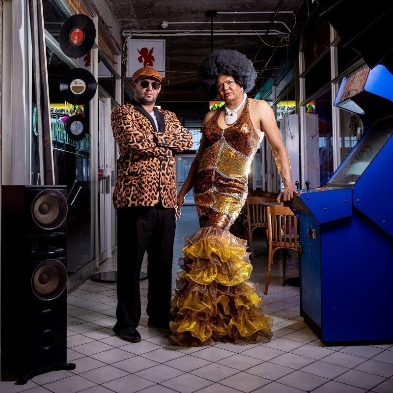 Constantina stands in a brown dress next to an arcade machine. With her is a member of the band Eduardo and the Night Train wearing an animal print jacket and black pants. 
Photographer Credit: Duane Preston