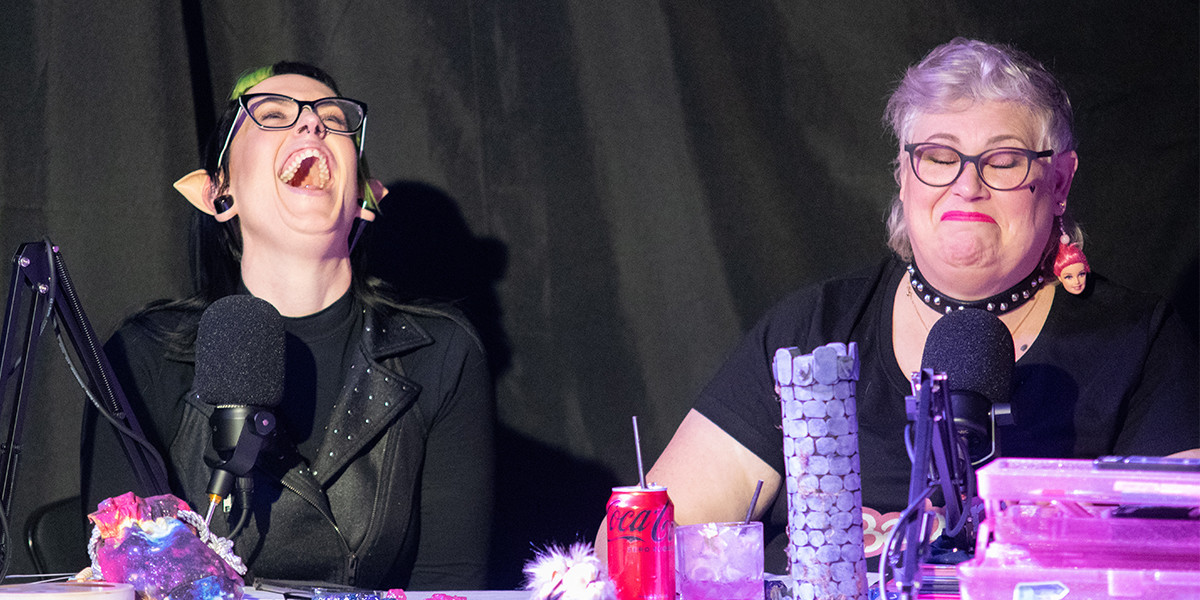 A picture of two femme people sitting at a D&D table. One of them has black glasses, is wearing a black leather jacket and fake long elf ears. She is throwing her head back in laughter. The other has short white hair, pink lipstick and a studded collar and she has a bemused look on her face.
