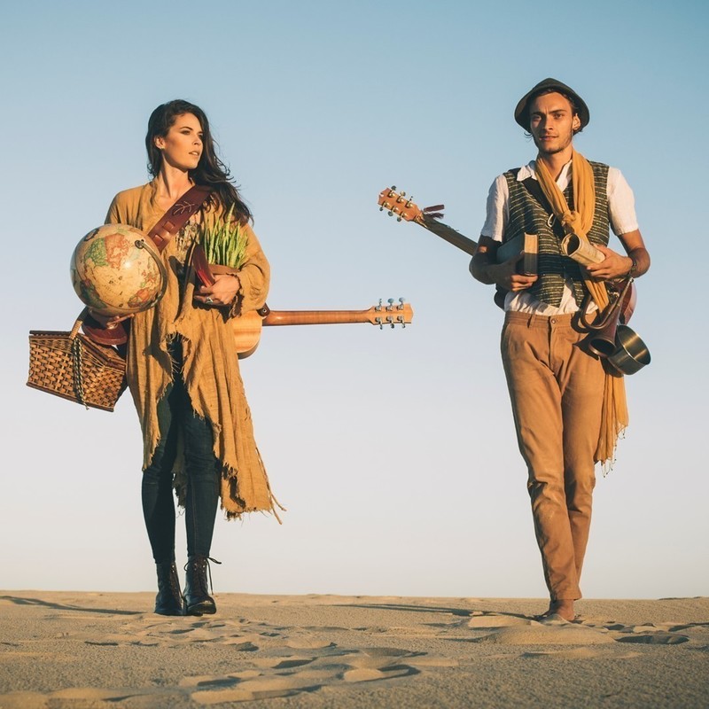 A photo of a man and a woman walking along the sand. The woman on the left of the image is holding a picnic basket, globe, and has a wooden musical instrument hanging on her back. The man on the right of the image is holding a book and a rolled up piece of paper with a wooden musical instrument hanging on his back.