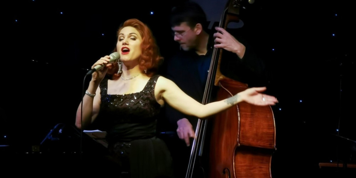 a singer with red hair sings into a microphone with her eyes closed and her arms outstreched. She is in a vintage black sequined gown. Behind her is a serious looking musician playing the double bass.