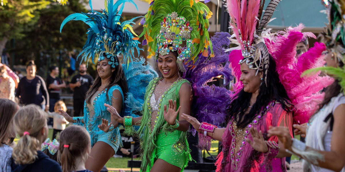 Brazilian style showgirls dancing with kids in an out door festival, cultural dance, samba dancers in colourful costumes, children learning Latin dancing, La Bomba dancers in Adelaide