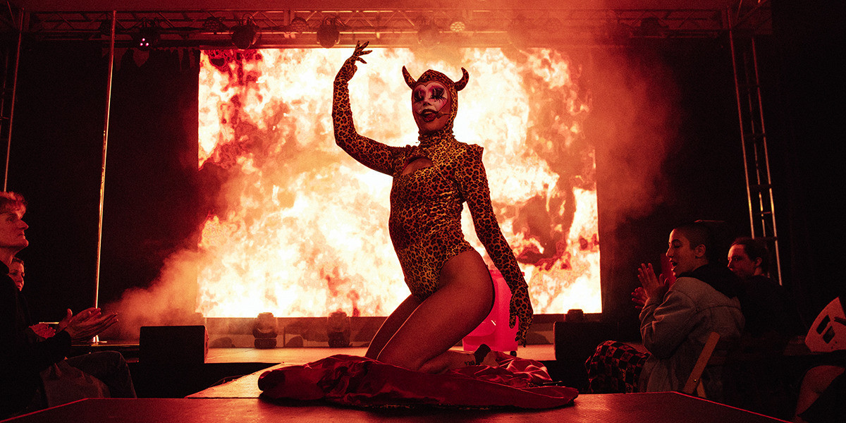 Themme Fatale, a performer with a heart shaped clown makeup, wears a devil horned leotard leopard costume with a big explosion in the background