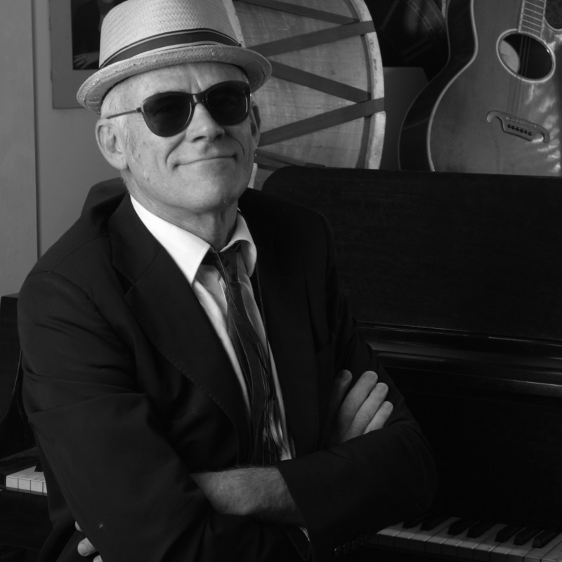 A greyscale photo of Stewart D'Arrietta, in a smart black suit, crisp white shirt and loosely tied tie, and a straw pork pie hat. 
He's sitting front on, with his arms folded across his chest, and you can just make out that his grand piano is next to him, on the left. Just behind him there's a kick drum, acoustic guitar and other assorted vintage musical paraphernalia propped up, as decoration. 
Stewart is wearing dark sunglasses and his chin is jutted out. He is smirking in a bemused way because he's about to play some of the politically cheeky songs of Randy Newman.