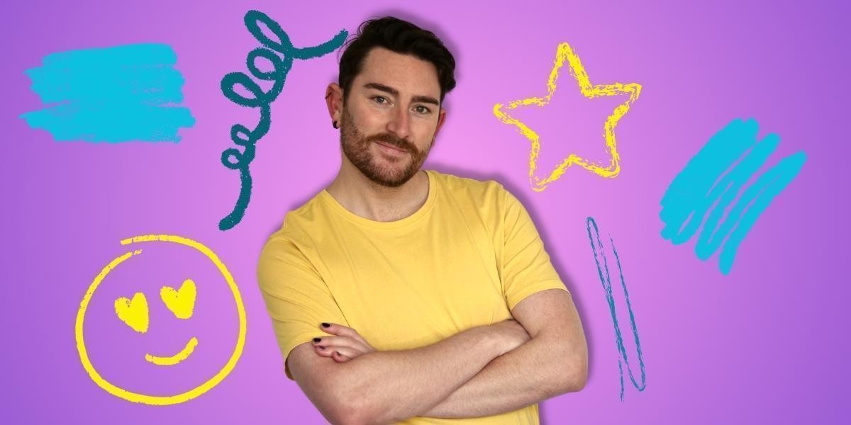 A man in a yellow shirt with his arms crossed smiling. With a purple background and an assortment of crayon drawing on the back wall: a star, a smiley face and squiggly lines.