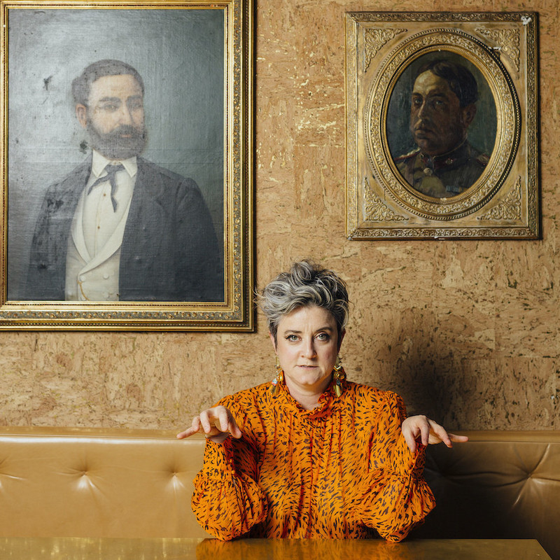 A performer sitting at a table in a bar dressed in an orange blouse with small black tiger stripes. She rests her elbows on the table and is holding both her hands up but dropped at the wrist like an animal claw. Her expression is neutral with a subtle glint in her eye. There are 2 old oil painting portraits of bearded men hung on the wall behind the performer.