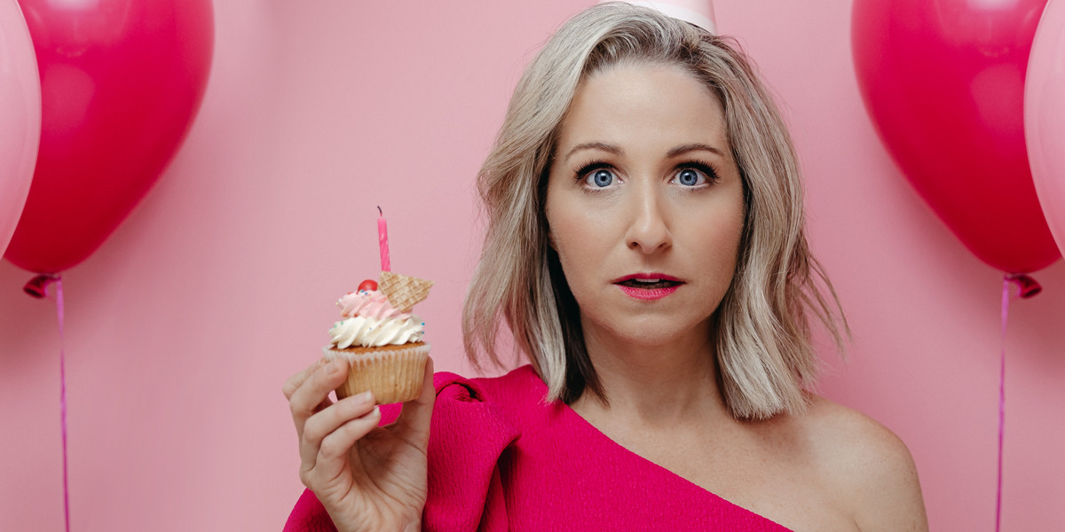 Jodie is wearing a hot pink party dress and party hat holding a birthday cake with a candle looking bewildered and perplexed. There is a pink party balloon in the background.