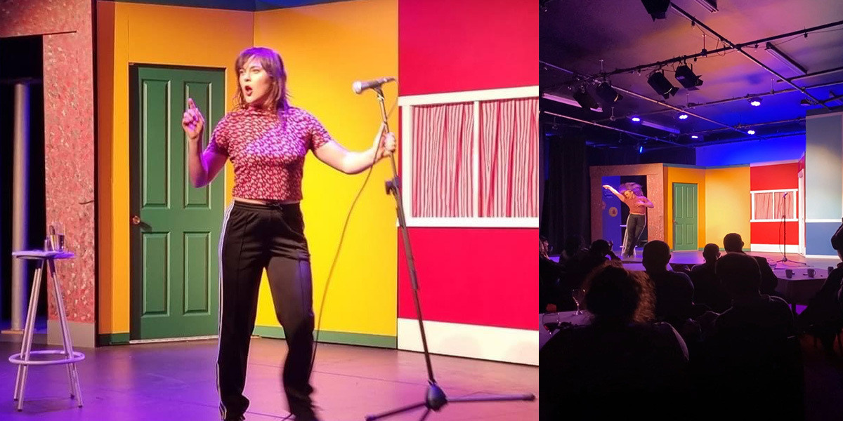 Joana Joy (NZ European Woman) onstage doing stand up comedy in a theatre with brightly coloured walls. Joana is holding the microphone stand and pointing at the audience. Joana wears a tight red t-shirt and black Adidas trackpants.