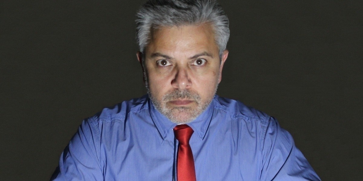 Man with grey hair wearing a blue buttoned shirt and red shiny tie. The man has a serious expression on his face.