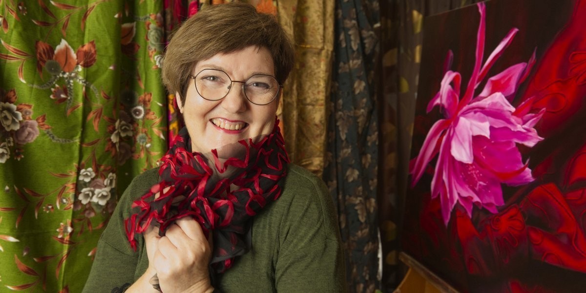 A dark-haired woman, wearing glasses, smiles. Artist Catherine Fitz-Gerald playfully poses in front of swathes of multi-coloured fabrics, a strip of red-on-black ribbon lace wound around her neck. To the right, on an angle, is her painting of a magenta flower on ruby red silk adorned with traceries of red cornelli work.