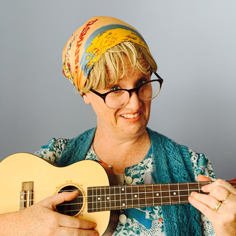 A photo of a woman wearing a colourful headscarf and an aquamarine coloured top with a confused expression on her face . She is holding a ukulele and is wearing glasses.