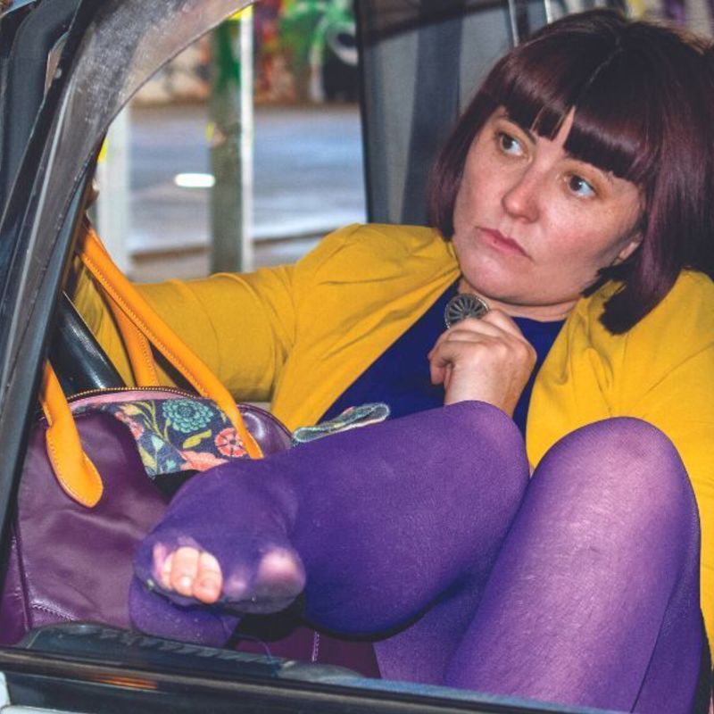 A woman sits in a white van, with purple tights and her legs hanging out of the front window.