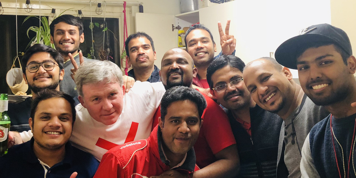 a group of about 12 men in a household kitchen pose in a happy party photo group. All of the faces but 1 seem to be South Asian
