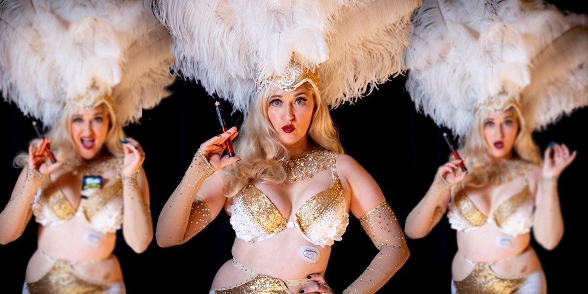 CANCELLED- The Diabetic Diva - Blonde woman in big white feathered head piece and gold and white costume holding an insulin needle. She has a diabetic device in her stomach.