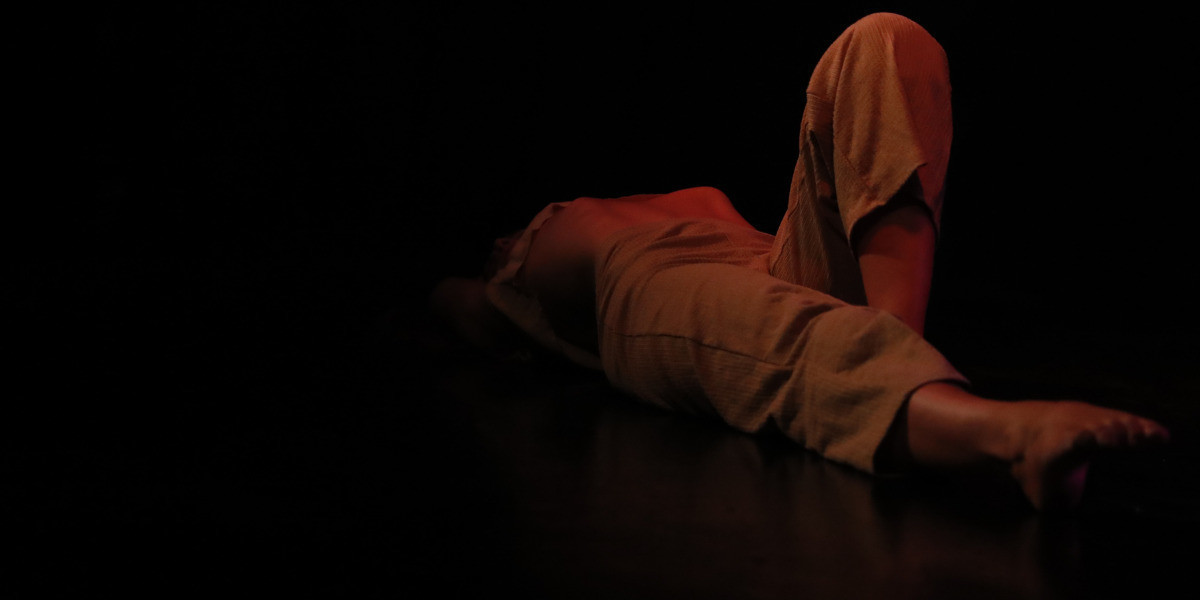 Woman laying on her back, background is black, the spotlight is moody and red in colour. You are unable to see her head as it fades into the background.
