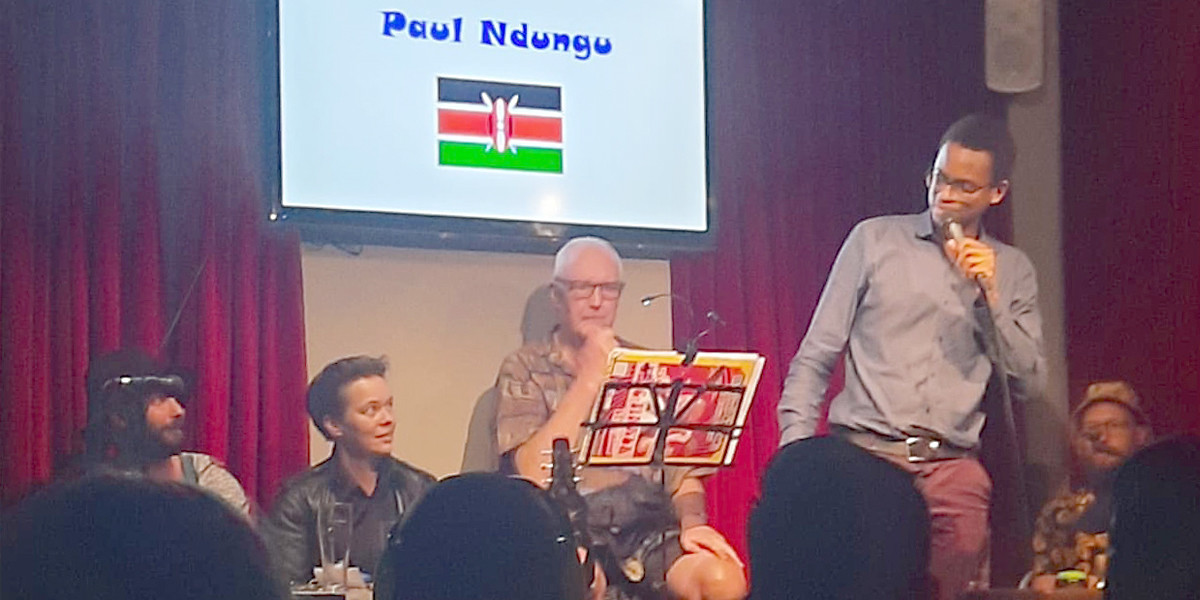Five comedians are on stage. On the left, seated, are Richard and Jess representing Team Adelaide. In the centre is MC Eric Tinker. On the right, for Team Rest of the World, are Paul (Kenya), standing and speaking, and Matt (USA), seated.