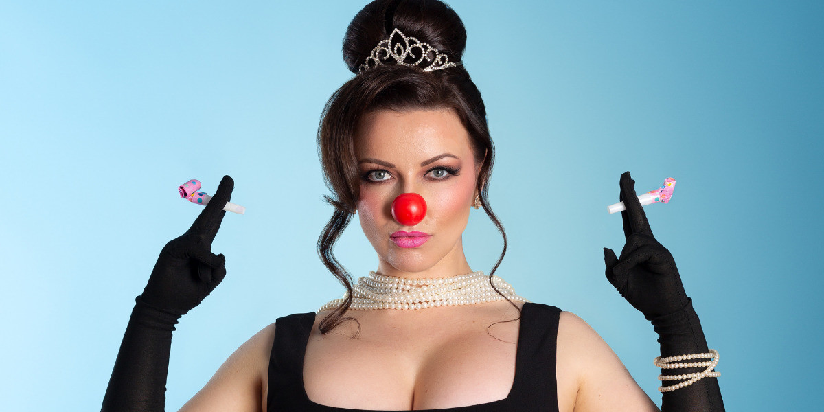 Close up of a woman wearing a red nose and holding two party poppers in each hand. She is also wearing black gloves.