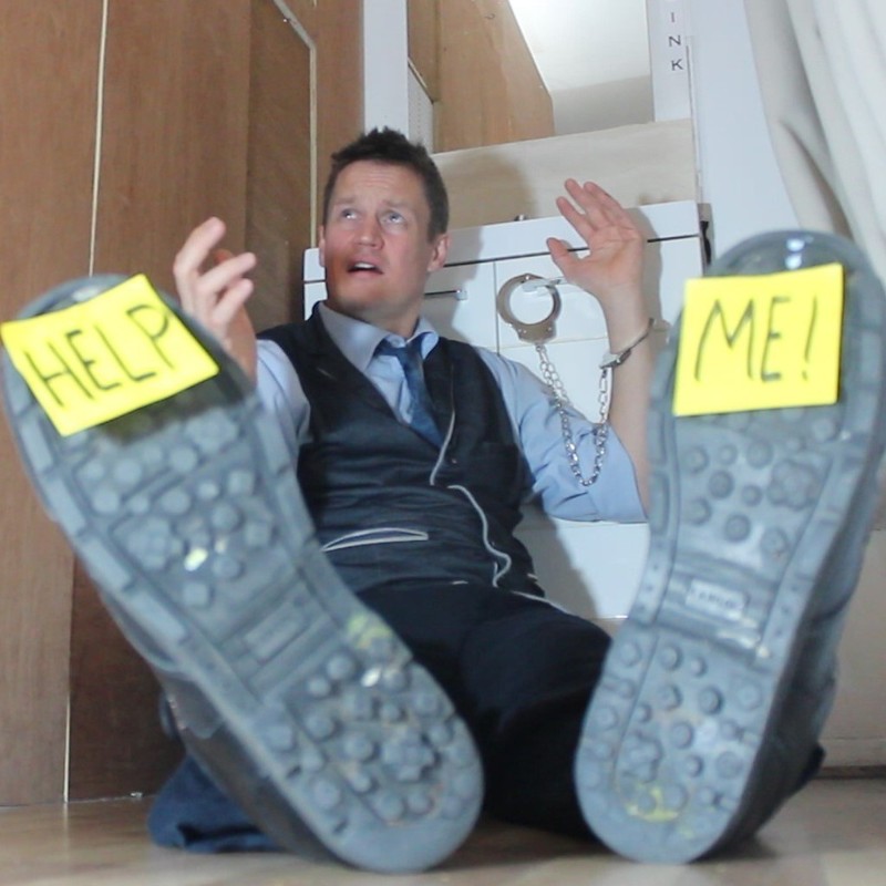 A man sitting on the ground with his legs out in front of him, he is handcuffed to a bathroom cabinet. There is a post it note on the sole of each of his shoes, the left says "help" the right says "me". He wears a blue tie, pale blue shirt and a waistcoat. His face has a pleading expression.