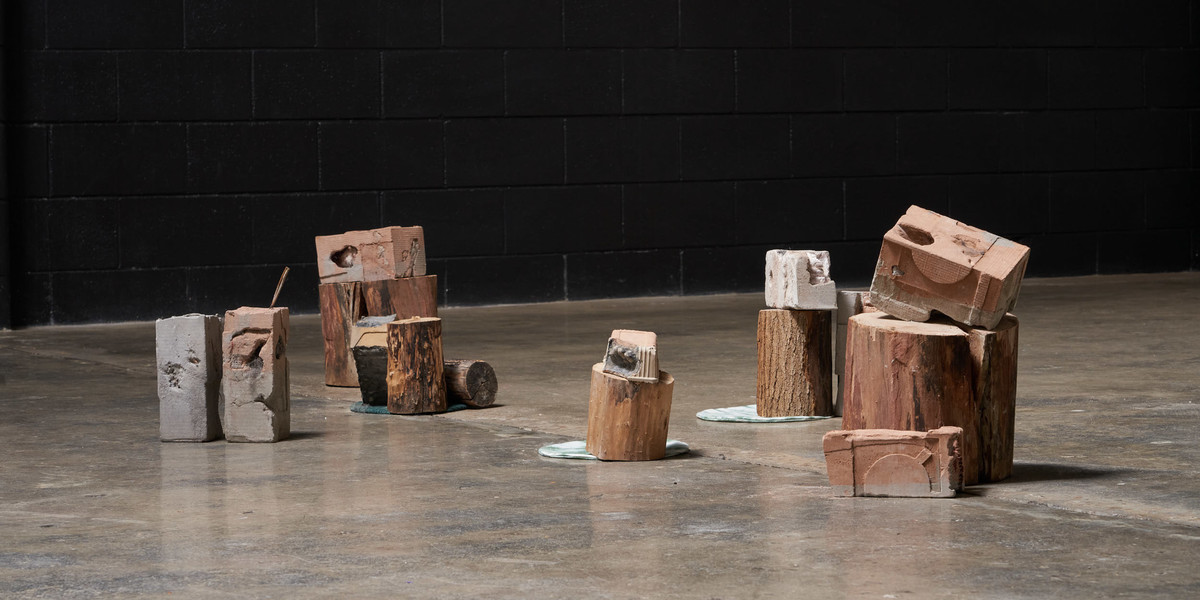 Meng Zhang, 'Container No 1 - 9', 2022, cast concrete, plaster, wood and hand-dyed fabric. Photography: Sam Roberts

Description: A collection of nine cast block forms with some resting on wooden logs. They sit on a concrete floor with a black wall in the background.