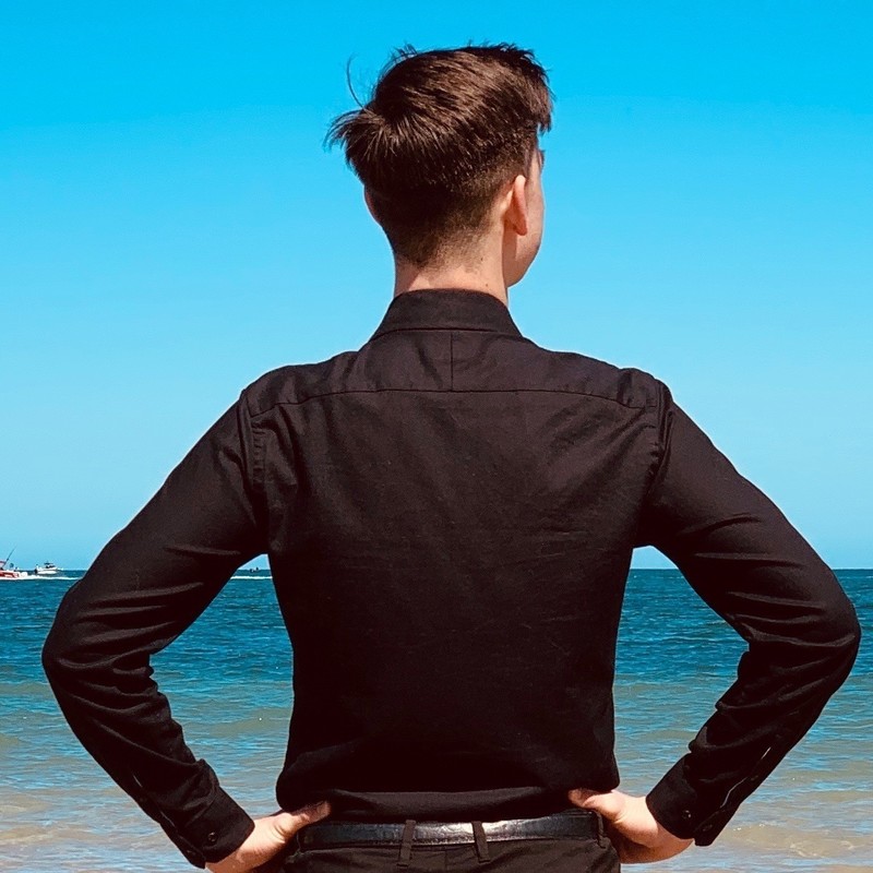 A man is standing with his back to the camera with his hands on his hips. He is wearing a long sleeve black shirt, he is standing on a beach.